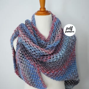 May include: A hand-knitted triangular shawl featuring a variegated yarn in shades of blue, grey, and pink. The accessory has an open, airy stitch pattern and is displayed on a mannequin. A circular white label with the text pdf pattern is visible.