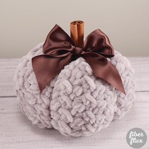 May include: A plush, gray pumpkin decoration with a brown satin bow and a cinnamon stick stem. The pumpkin is made of a soft, textured material, perfect for fall decor. The Fiber Flux logo is visible in the bottom right corner.