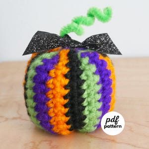 May include: A handmade crochet pumpkin in Halloween colors: green, purple, orange, and black. It has a black glitter bow and a green pipe cleaner stem. The pumpkin is a round shape and is sitting on a wooden surface. The image includes the text "pdf pattern".