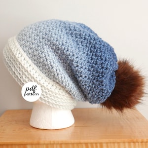 Op de afbeelding: Een handgehaakte muts in blauw- en wittinten, afgewerkt met een bruine imitatiebont pompon. De muts wordt getoond op een witte mannequinhoofd. De tekst "pdf pattern" staat op de voorkant.