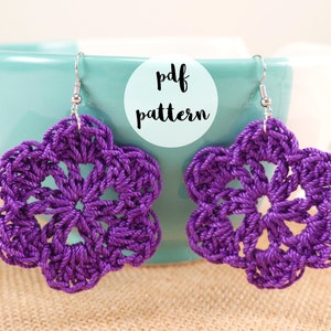 以下が含まれることがあります： シルバーのフックが付いた紫色のクロシェフラワーのイヤリング。イヤリングはターコイズブルーのボウルからぶら下がっています。白い円に「pdf pattern」というテキストが書かれています。