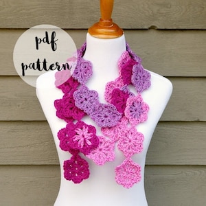 Peut inclure: Une écharpe en crochet de fleurs dans des tons de rose et de violet. L'écharpe est présentée sur un mannequin blanc. Le texte "pdf pattern" est dans un cercle au-dessus de l'écharpe. Le texte "fiber flux" est en dessous de l'écharpe.