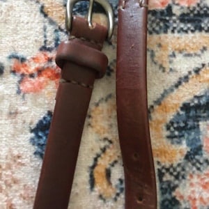 Può includere: Primo piano di una cintura in pelle marrone con fibbia color argento. La cintura è di un ricco colore marrone scuro e sembra essere realizzata in pelle di alta qualità. La fibbia è un design semplice e classico. La cintura è probabilmente per pantaloni o gonne.