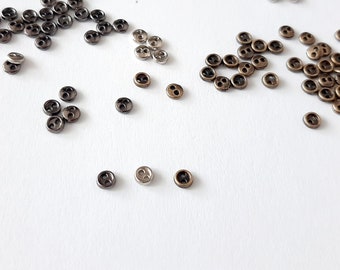 May include: A collection of small, round, metal buttons in silver, black, and bronze colors. The buttons are scattered on a white surface.