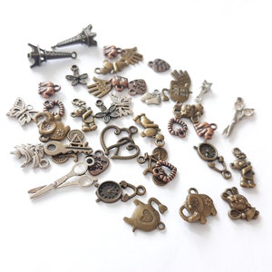 May include: Assortment of metal charms in various shapes and designs, including hearts, teapots, butterflies, scissors, and a small Eiffel Tower. The charms are in shades of gold, silver, and copper.