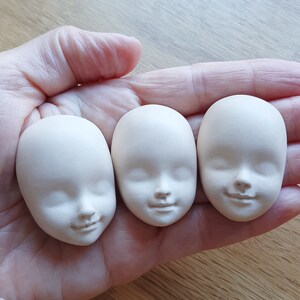 May include: Three unfinished white porcelain doll heads with closed eyes and sculpted facial features. The heads are arranged in a row on a hand.