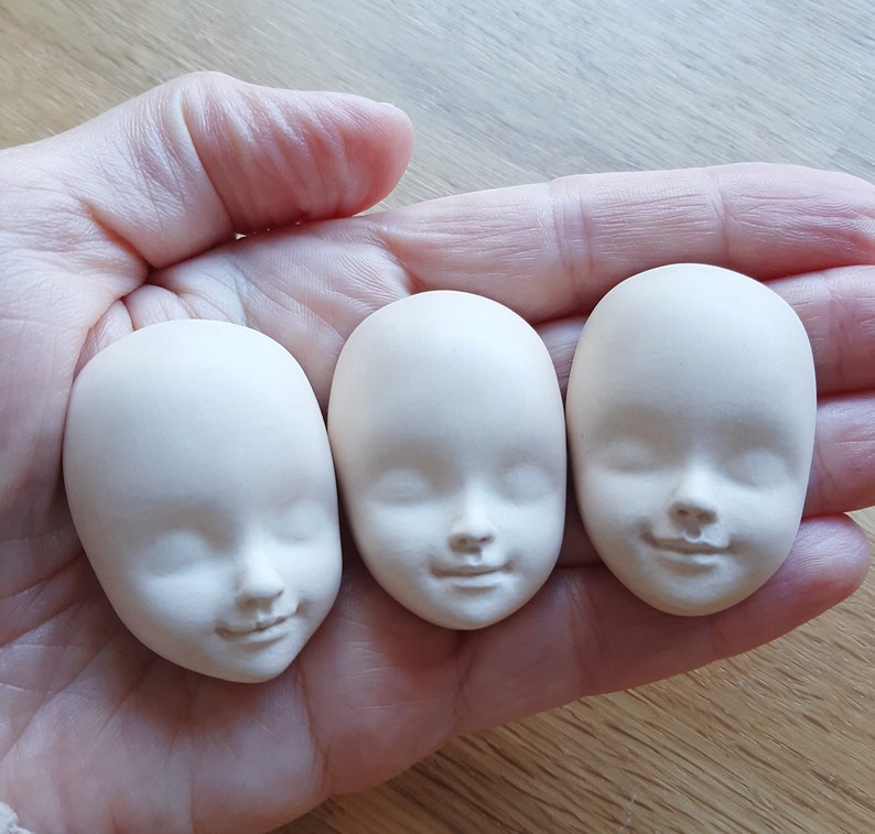 May include: Three unfinished white porcelain doll heads with closed eyes and sculpted facial features. The heads are arranged in a row on a hand.