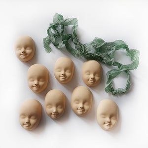 May include: Eight unpainted plaster doll faces with a green fabric ribbon. The faces have molded features, including eyes, noses, and mouths.
