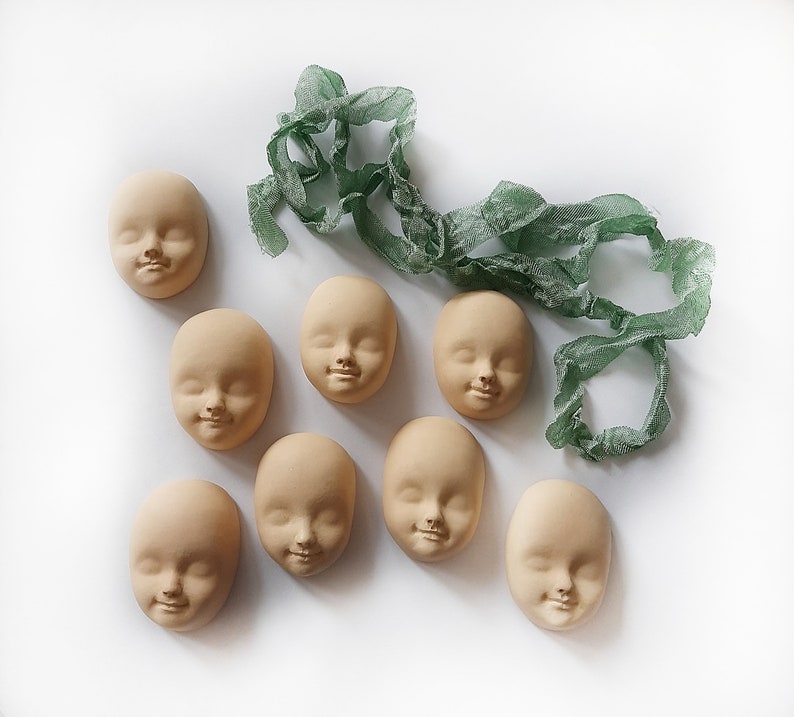 May include: Eight unpainted plaster doll faces with a green fabric ribbon. The faces have molded features, including eyes, noses, and mouths.