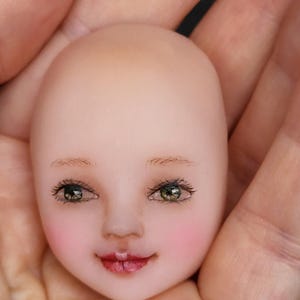 Op de afbeelding: Een porseleinen poppenhoofd met geschilderde groene ogen, een roze blos en een glimlach met gesloten mond.