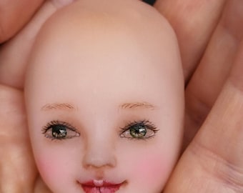 May include: A porcelain doll head with painted green eyes, a pink blush, and a closed-mouth smile.