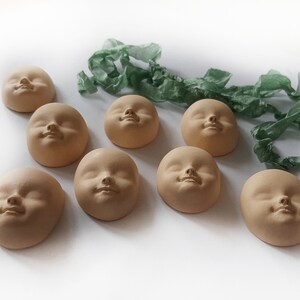 May include: Eight small, unglazed ceramic sculptures of faces, each with a different expression, arranged on a white surface. The sculptures are all the same size and shape, and they are all made of a light brown clay.