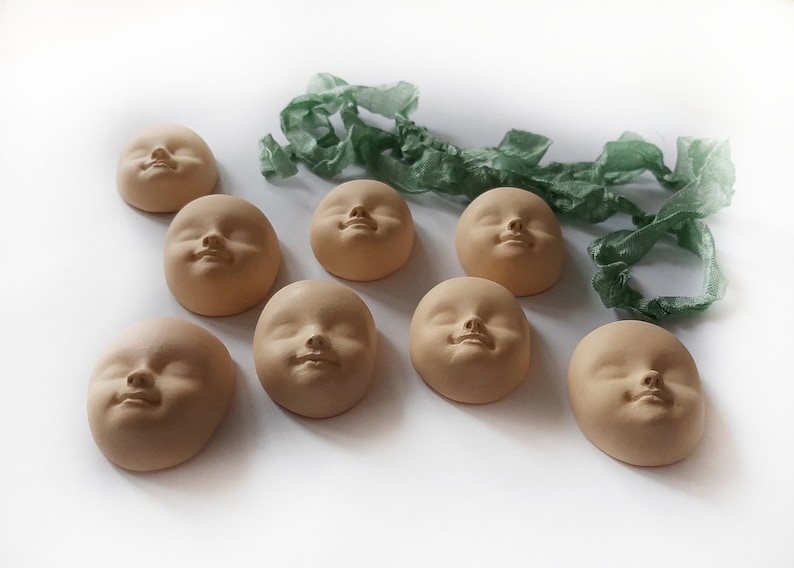 May include: Eight small, unglazed ceramic sculptures of faces, each with a different expression, arranged on a white surface. The sculptures are all the same size and shape, and they are all made of a light brown clay.