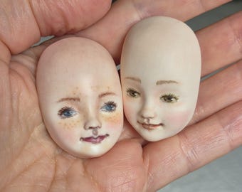 May include: Two oval-shaped doll heads with hand-painted facial features. The left head features blue eyes, freckles, and a pink mouth. The right head features brown eyes, light eyebrows, and a soft pink blush on the cheeks. Both have smooth skin.