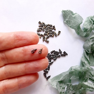 May include: A close-up of two small, gold-toned metal triangle-shaped toggles with two holes each. The toggles are on a finger, and there are many more toggles scattered on a white surface.