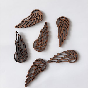 May include: Six wooden angel wing cutouts in various sizes and orientations. The wings are a warm brown color with a detailed, openwork design. The cutouts are arranged on a white background.