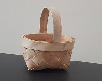 Op de afbeelding: Een klein, geweven mandje met een natuurlijke houten afwerking en een enkele handgreep. Het mandje is leeg en staat op een donker oppervlak.