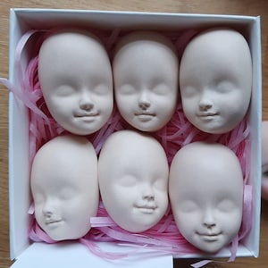 May include: Six white porcelain doll heads with closed eyes and painted features. The heads are arranged in a box with pink tissue paper.