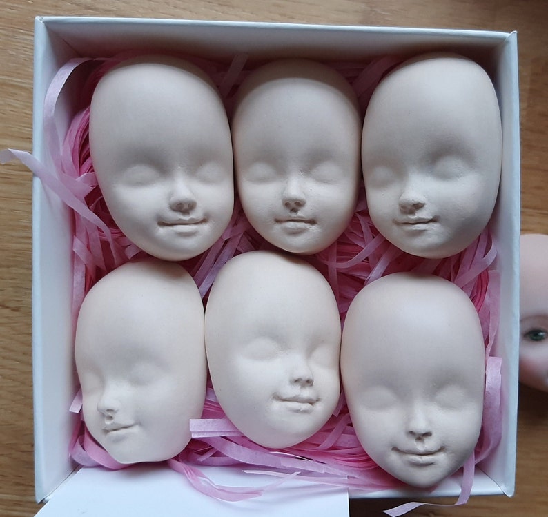 May include: Six white porcelain doll heads with closed eyes and painted features. The heads are arranged in a box with pink tissue paper.