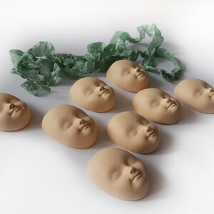 May include: Eight ceramic baby face sculptures in a light brown color, arranged in a row on a white surface. A green ribbon is in the background.