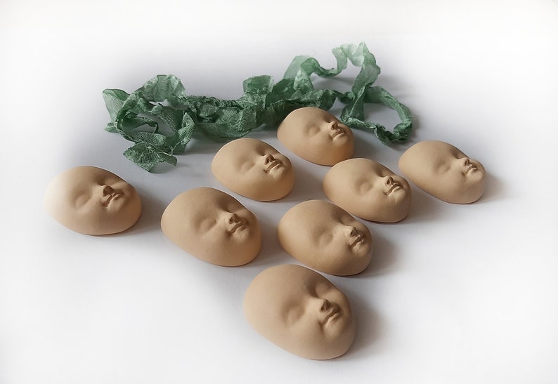 May include: Eight ceramic baby face sculptures in a light brown color, arranged in a row on a white surface. A green ribbon is in the background.