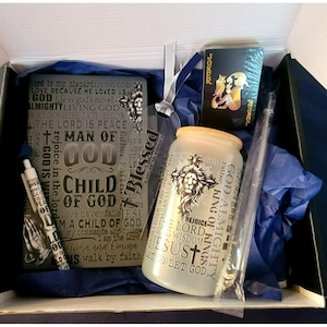 Puede incluir: Un conjunto de regalo con una variedad de artículos religiosos. El conjunto incluye una libreta con un diseño en blanco y negro con las palabras "Blessed" y "Child of God", un frasco de vidrio con tapa de madera y un diseño de león con las palabras "King of Kings" y "God Almighty", un bolígrafo blanco y negro con las palabras "Living God" impresas en él, y una baraja de cartas negra con un diseño de joker dorado.