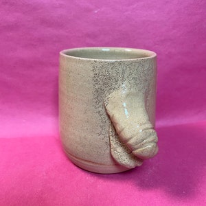 May include: A beige ceramic mug with a unique handle shaped like a stylized appendage. The mug has a speckled texture and sits against a bright pink background. The mug is cylindrical and appears handmade.