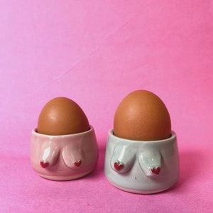 Può includere: Due portauova in ceramica, uno rosa e uno verde-azzurro, ciascuno a forma di busto con dettagli a forma di cuore rosso. Ogni tazza contiene un uovo marrone. Lo sfondo è rosa.