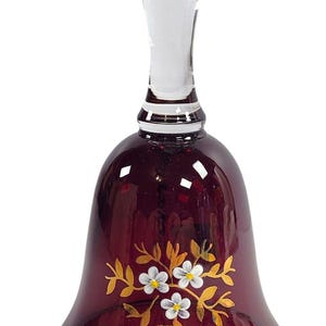 Puede incluir: Una campana de cristal rojo rubí con un asa de cristal transparente. La campana presenta un borde dorado y un diseño floral con flores blancas y hojas doradas. Un pequeño punto negro está en la parte superior del asa. Artículo decorativo.