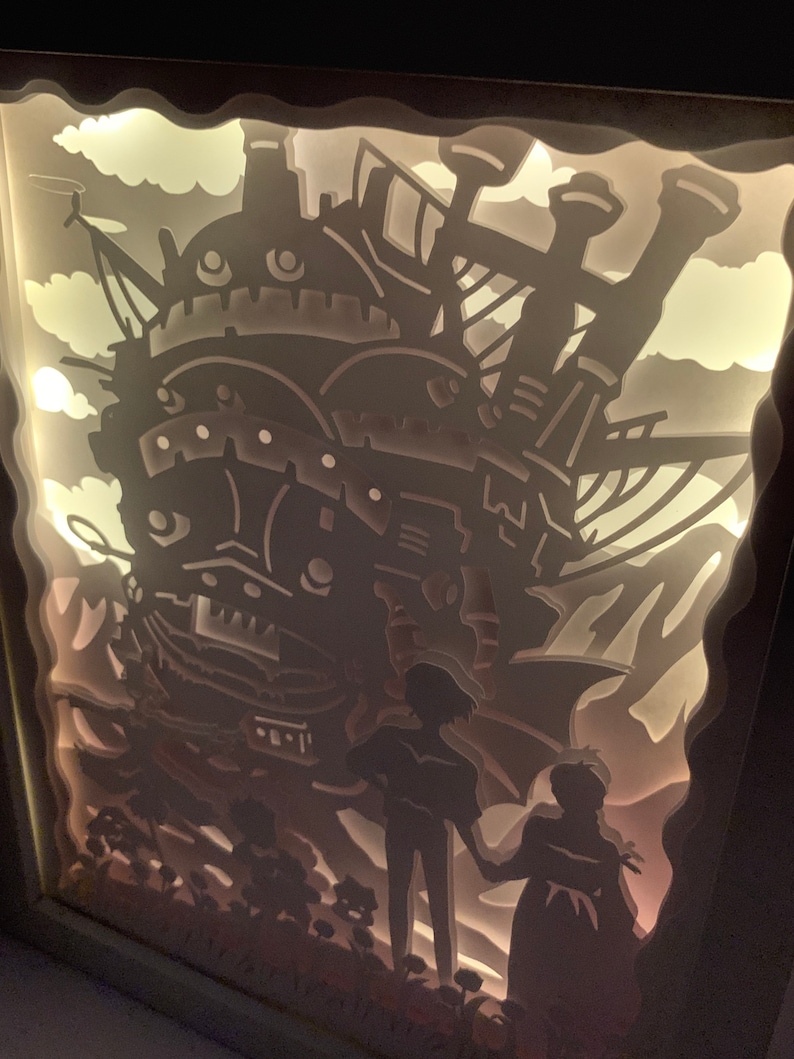 May include: A white papercraft light-up display of a flying airship with two figures standing in front of it. The airship is detailed with windows and a large chimney. The figures are silhouetted against the airship.