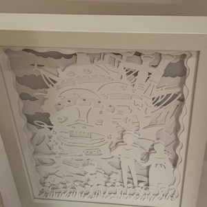 May include: White paper cut-out art framed in a white frame. The art depicts a flying house with two figures standing in front of it. The house is detailed with windows and a chimney. The figures are holding hands and are standing in a field of flowers.