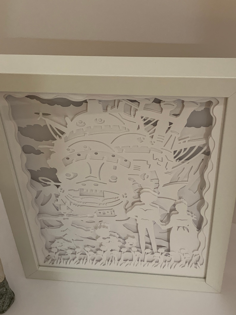 May include: White paper cut-out art framed in a white frame. The art depicts a flying house with two figures standing in front of it. The house is detailed with windows and a chimney. The figures are holding hands and are standing in a field of flowers.