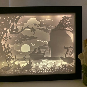 Könnte beinhalten: Ein gerahmtes, beleuchtetes Papierkunstwerk, das eine ruhige Seeszene darstellt. Das Kunstwerk zeigt ein Boot mit zwei Figuren, Schwänen und einem Vollmond. Der Rahmen ist schwarz, und das Kunstwerk wird von hinten beleuchtet, wodurch ein Silhouetteffekt entsteht.