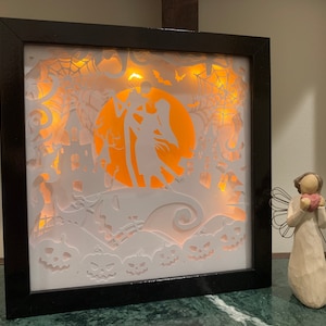May include: A black framed shadow box with a white paper cutout design of Jack Skellington and Sally from the Nightmare Before Christmas. The design includes a spiderweb, bats, and pumpkins. The box is lit from behind with warm white lights.
