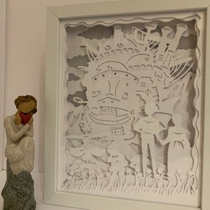 May include: White paper cut-out art framed in a white frame. The art depicts a flying castle with a couple standing in front of it. The castle is detailed with windows and chimneys. The couple are holding hands and looking at each other. The background is a sky with clouds.