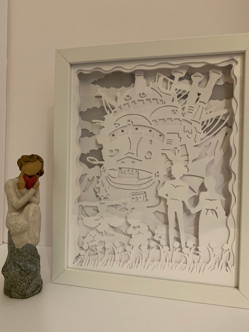 May include: White paper cut-out art framed in a white frame. The art depicts a flying castle with a couple standing in front of it. The castle is detailed with windows and chimneys. The couple are holding hands and looking at each other. The background is a sky with clouds.