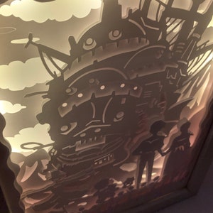 May include: A white papercraft lamp featuring a silhouette of a flying machine, two figures, and clouds. The lamp is lit from behind, creating a glowing effect.