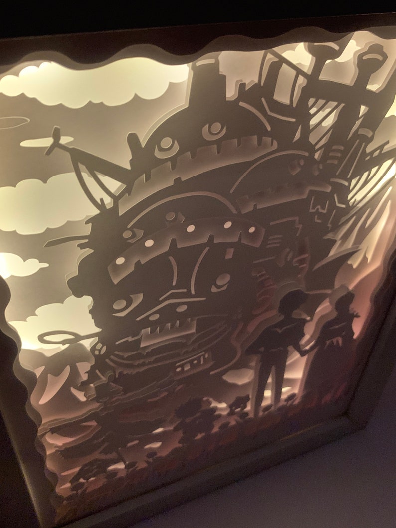 May include: A white papercraft lamp featuring a silhouette of a flying machine, two figures, and clouds. The lamp is lit from behind, creating a glowing effect.