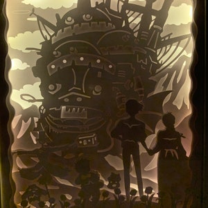 May include: A papercraft light-up display featuring a silhouette of a flying machine from the animated film Howl's Moving Castle. The machine is surrounded by clouds and flowers, and two figures stand in front of it.