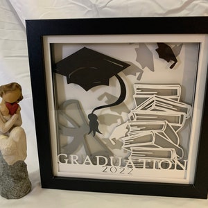 Könnte beinhalten: Eine schwarze, gerahmte Schattenbox mit weißem Hintergrund. Die Schattenbox zeigt eine schwarze Graduation Cap, einen Stapel Bücher und den Text "GRADUATION 2022".