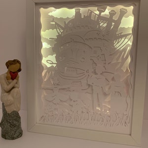 May include: A white framed light-up shadow box featuring a white paper cut-out design of a flying castle with clouds and grass. The castle is illuminated from behind, creating a glowing effect.