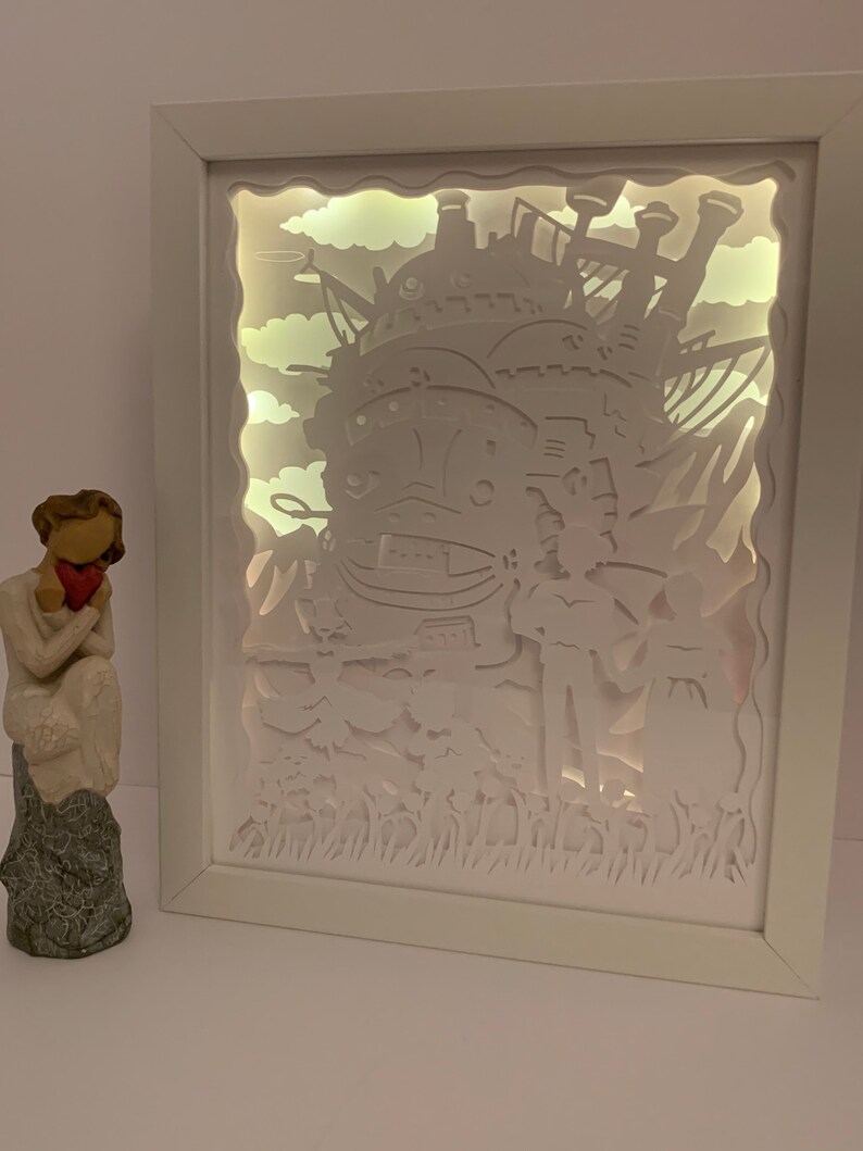 May include: A white framed light-up shadow box featuring a white paper cut-out design of a flying castle with clouds and grass. The castle is illuminated from behind, creating a glowing effect.