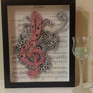 May include: A framed piece of art featuring a red treble clef with black and white floral designs on a sheet of music. The music is titled "Ave Maria" and is transcribed by Franz Schubert.