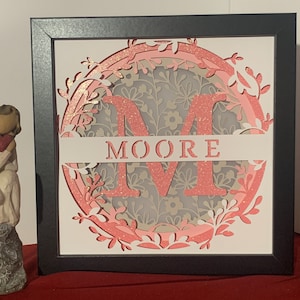 May include: A framed pink and white paper cut-out design with the letter M and the word MOORE. The design is surrounded by a floral pattern.