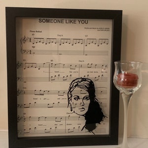 May include: A framed print of sheet music for the song "Someone Like You" by Adele. The sheet music is in black and white and features a silhouette of Adele's face.