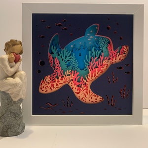 May include: A white frame with a blue background featuring a paper cut-out of a sea turtle with coral and seaweed inside. The turtle is swimming upwards with its head facing the left side of the frame.