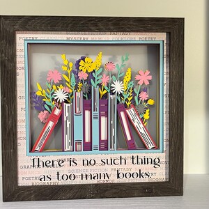 May include: A framed paper art piece featuring a colorful arrangement of paper-cut books and flowers. The artwork includes the text "There is no such thing as too many books" and is set against a background of book-related words.