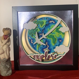 Op de afbeelding: Een ingelijst papieren kunstwerk met een 3D-laagontwerp van een wereldbol met vliegtuigen die eromheen vliegen. De wereldbol is omgeven door wolken en het woord "explore" is in een decoratief lettertype onder de wereldbol geschreven.