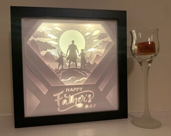 Könnte beinhalten: Eine schwarze Schattenbox mit einem weißen Papier-Ausschnitt-Design. Das Design zeigt eine Silhouette eines Vaters und zweier Kinder, die auf einem Berggipfel mit einem Vollmond im Hintergrund stehen. Der Text "Happy Father's Day" befindet sich am unteren Rand des Designs.