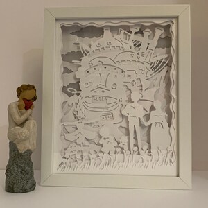 May include: A white paper cut-out art piece in a white frame depicting a flying castle with two figures standing in front of it. The castle is surrounded by clouds and flowers.